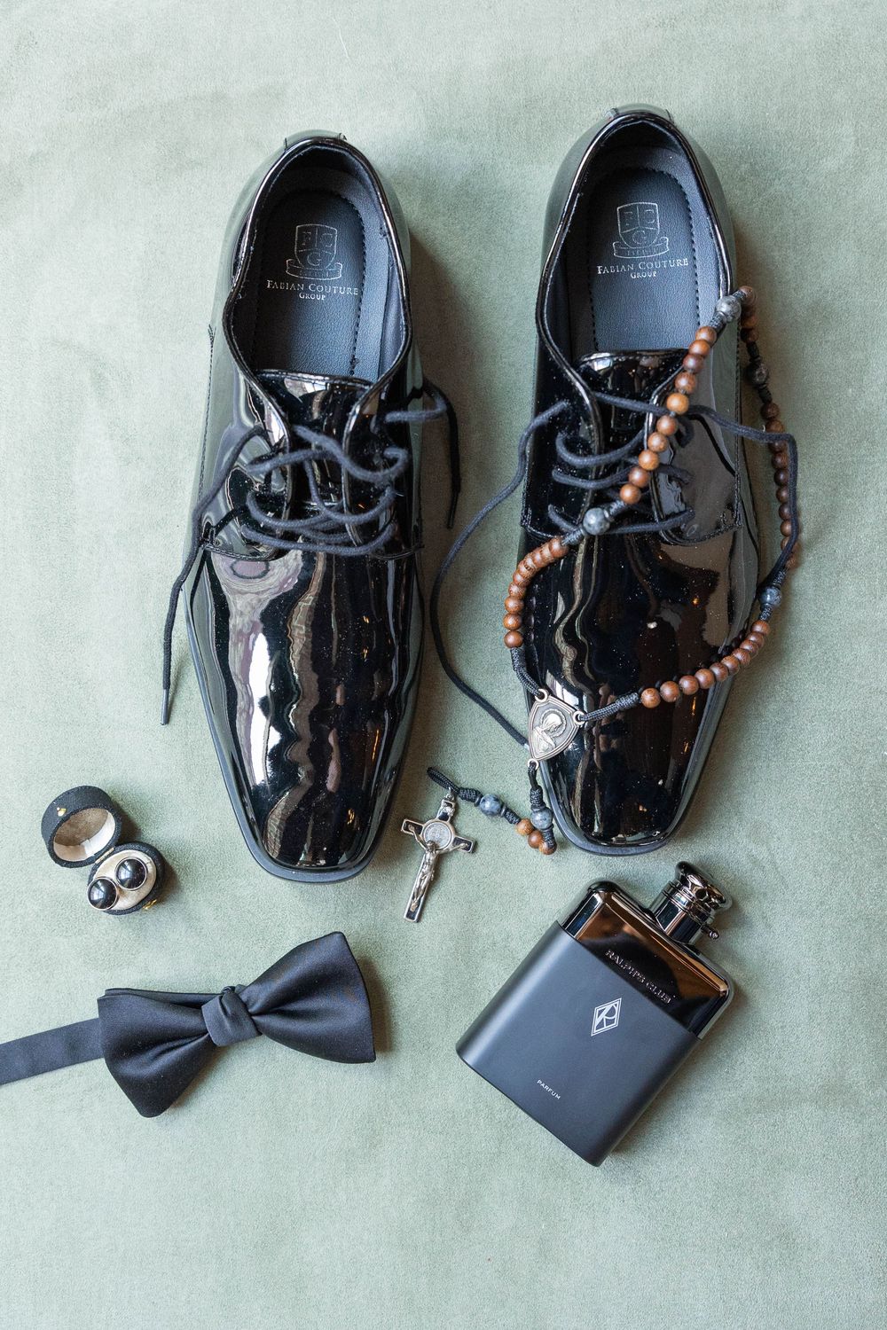 Groom's wedding accessories including black patent leather dress shoes, bow tie, rosary beads, cufflinks and flask on green background.