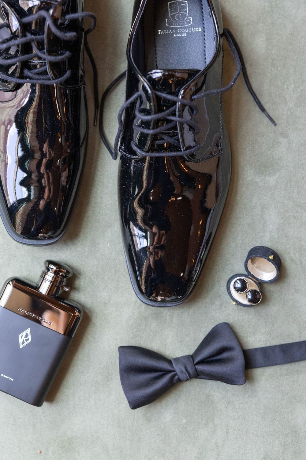 Patent leather dress shoes arranged with black bow tie, cologne bottle and cufflinks on gray background.