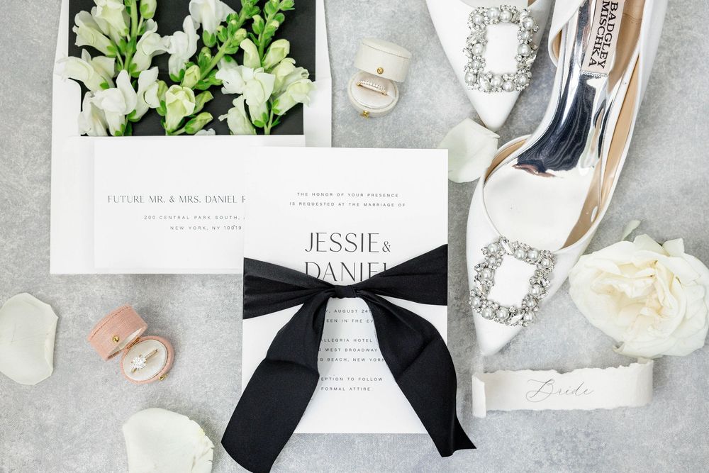 White wedding invitation with black bow ribbon surrounded by floral elements and bridal accessories on gray surface.