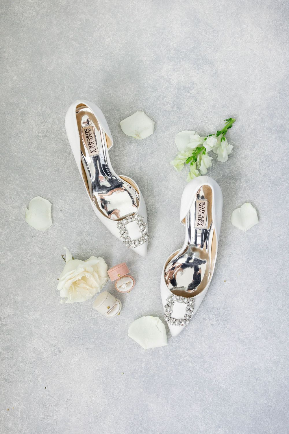 Elegant silver bridal shoes with crystal embellishments styled with white roses on light grey textured background.