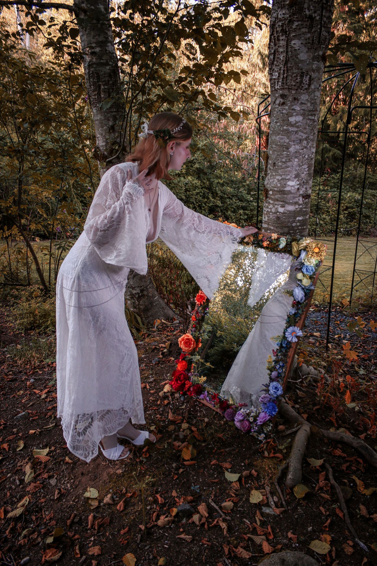 A figure in a flowing white vintage dress arranges flowers around an ornate mirror in a mystical forest at dusk.