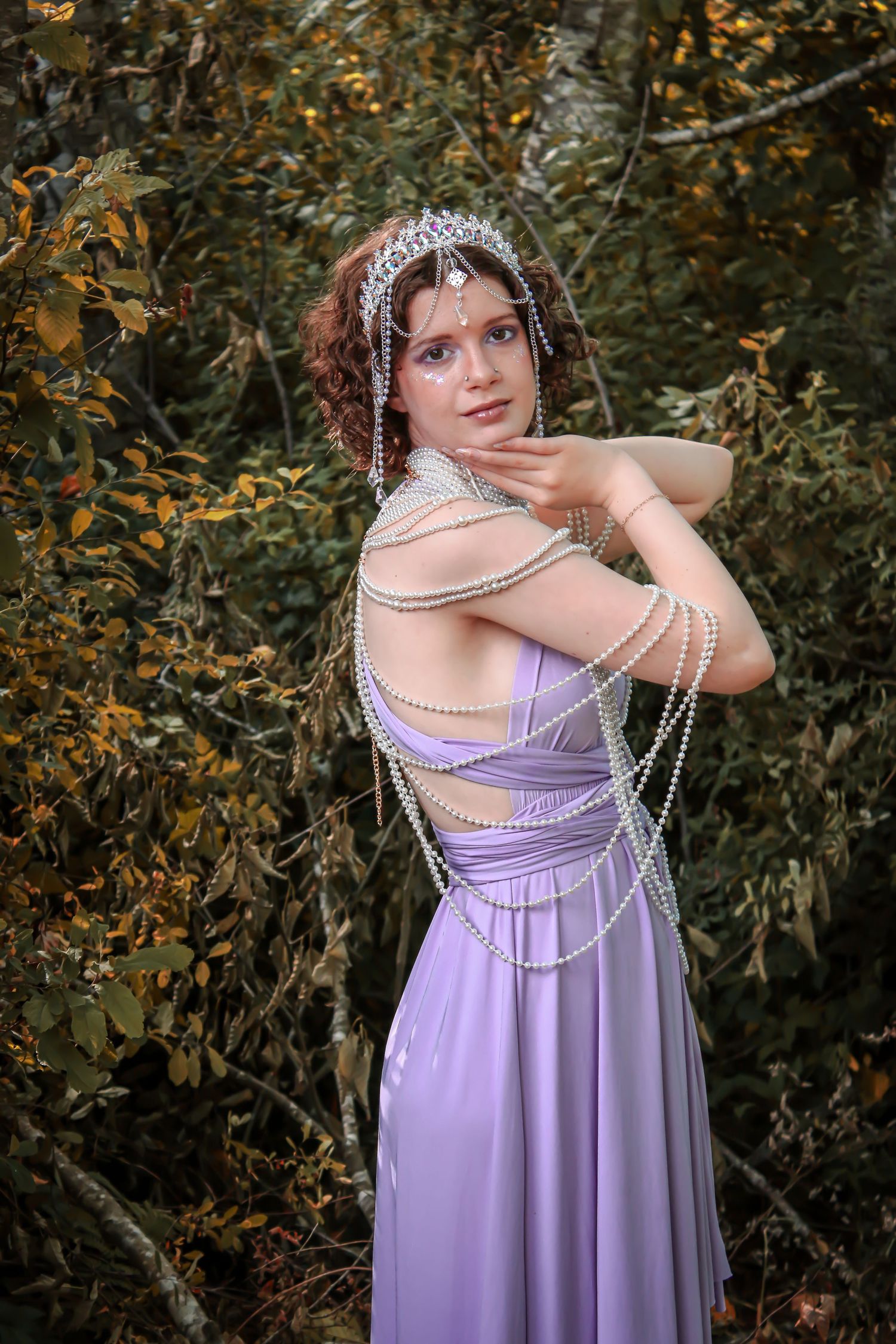 A person in a lilac dress with pearl strands draped across their shoulders poses elegantly in a natural woodland setting.