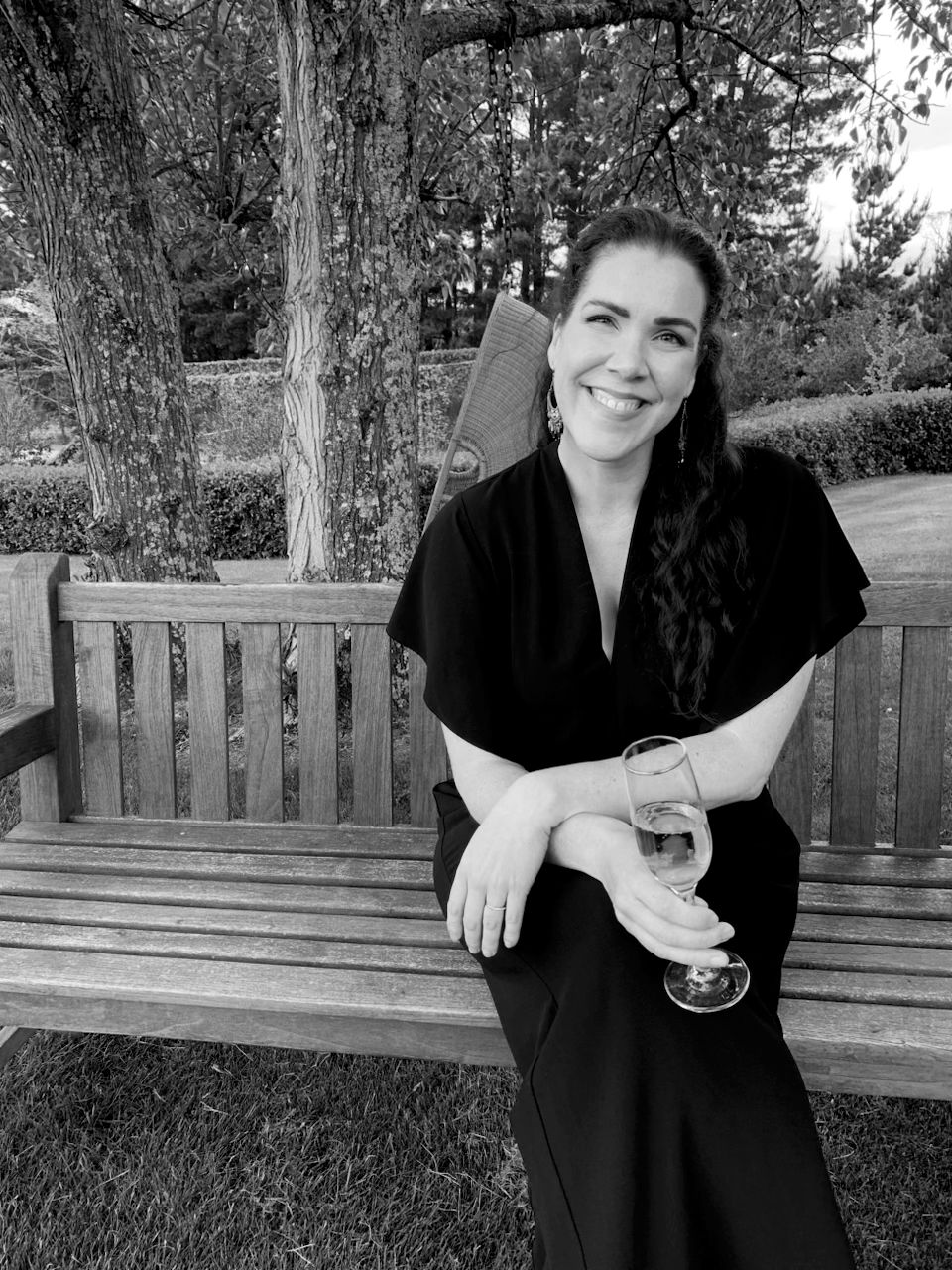 A person in a black dress sits smiling on a wooden bench in an outdoor garden setting, captured in black and white.