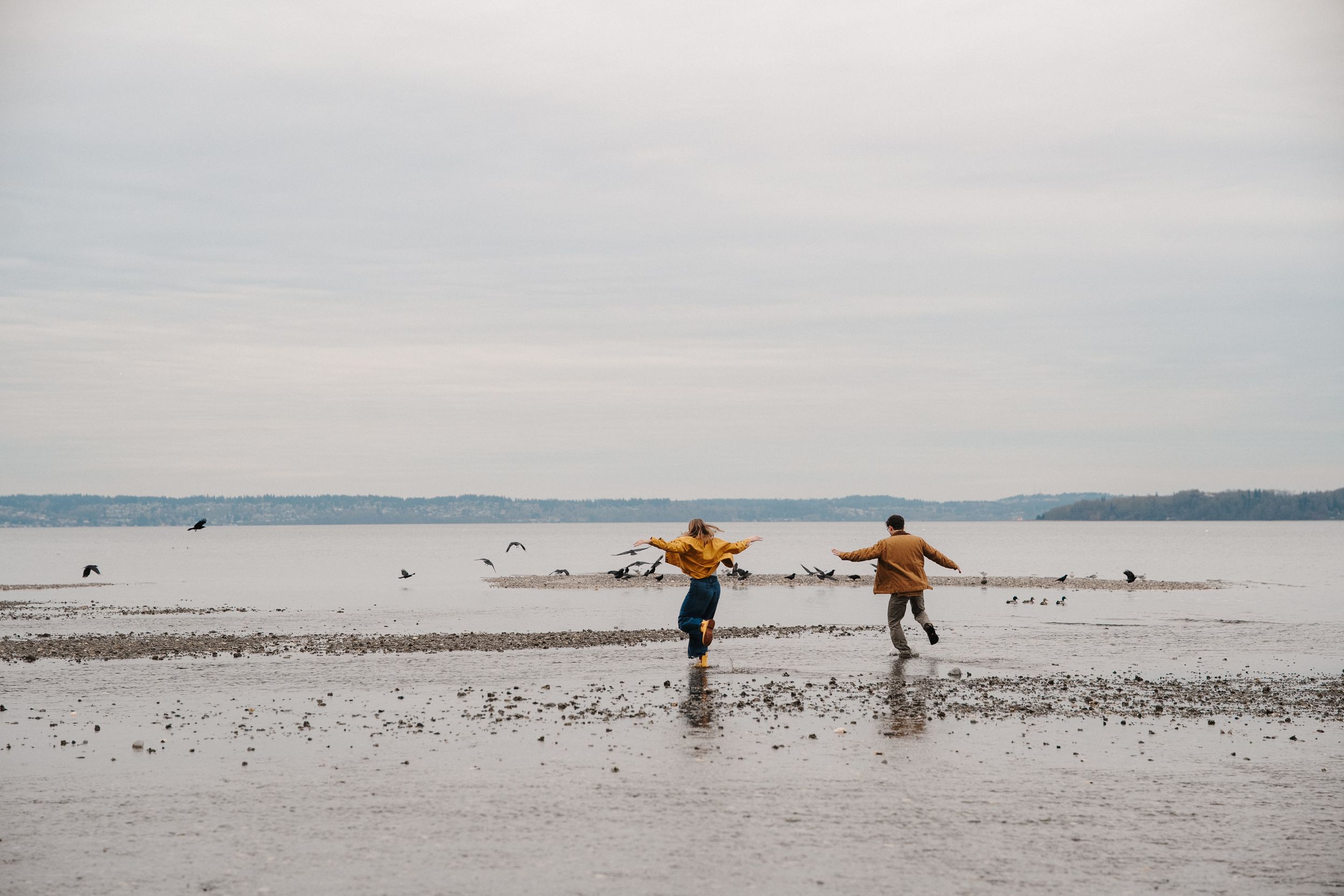 Two children in yellow raincoats play on a wet sandy beach on an overcast day, arms outstretched with joy.