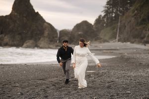 A couple runs joyfully on a moody, rocky beach surrounded by dramatic coastal cliffs on an overcast day.