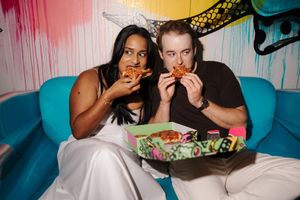 Two friends sit on a teal sofa eating chicken wings together in a colorful, graffiti-decorated room.