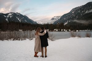 Couple embracing in snowy winter landscape with mountain lake and pine forest backdrop at dusk.