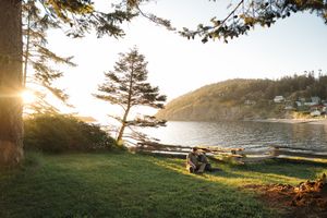Sun setting over a peaceful bay with silhouetted trees and a couple relaxing on grass by the water's edge.