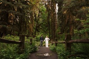 A figure in white walks along a scenic forest path surrounded by towering trees and lush greenery.
