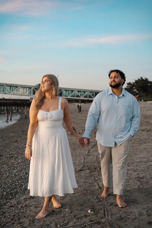 Couple walking barefoot on beach at sunset holding hands with bridge in background under blue sky.