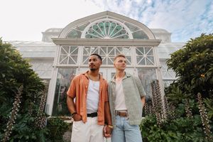 Two men standing in front of a grand glass greenhouse surrounded by lush green plants on a cloudy day.