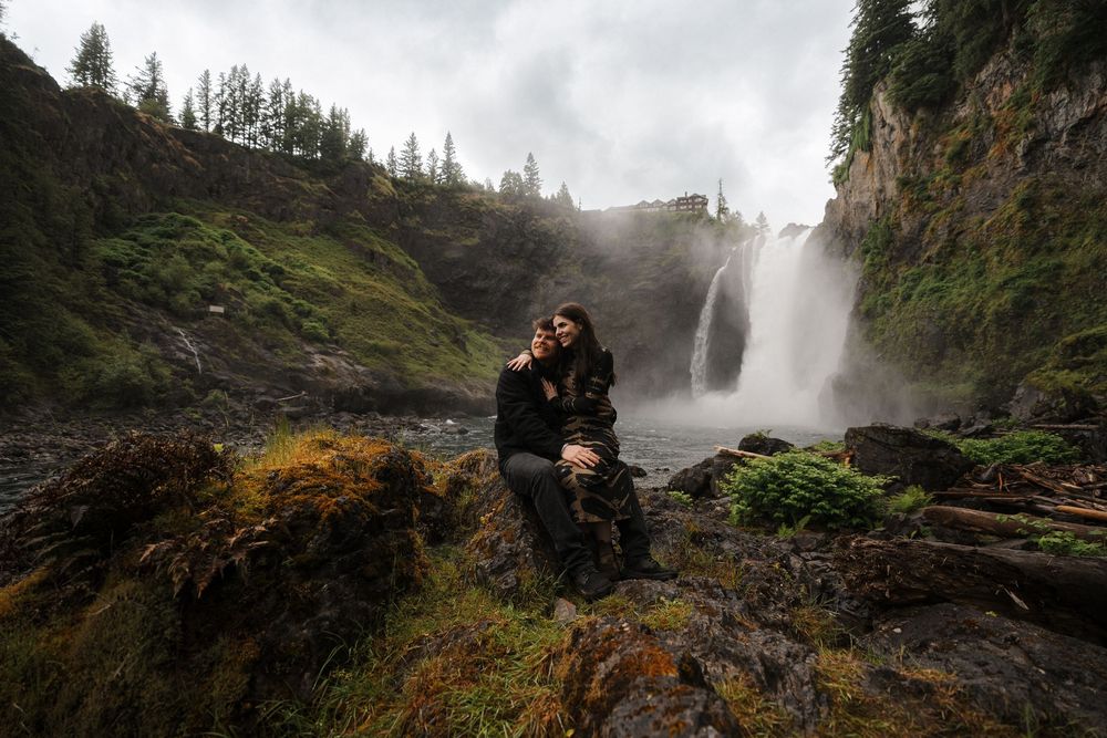 Couple embracing on rocks before a majestic waterfall surrounded by lush green cliffs on a misty day.