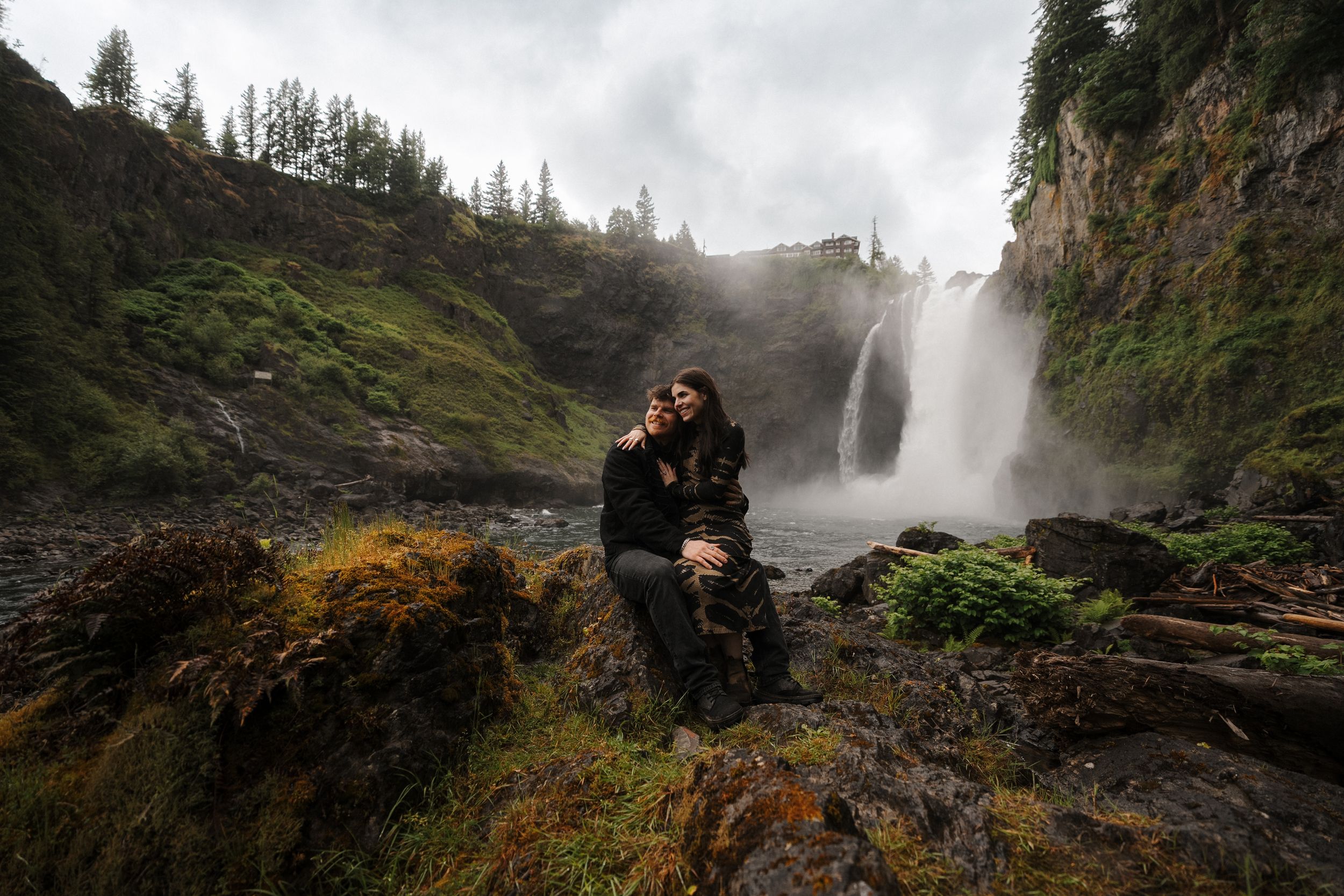 Couple embracing on rocks before a majestic waterfall surrounded by lush green cliffs on a misty day.