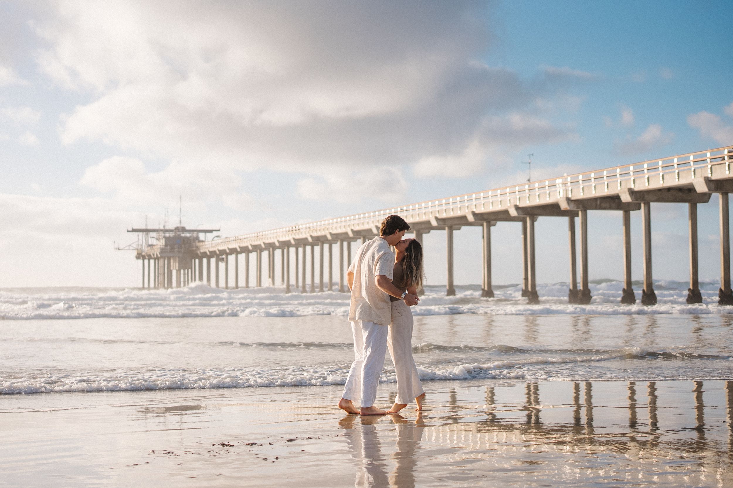 A couple in white clothing embraces near a wooden pier extending into the ocean at golden hour.