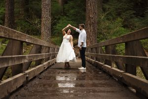 A couple in wedding attire dances together on a rustic wooden bridge surrounded by tall pine trees.