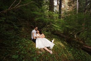 A couple in wedding attire sits together among lush green forest foliage during an outdoor photography session.