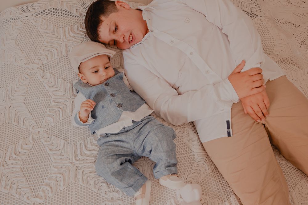 Person in white shirt holds and plays with baby wearing gray outfit against neutral background.