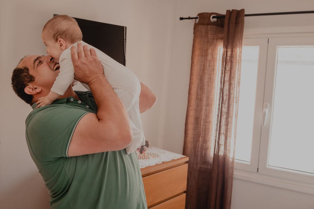 A tender moment captured as a parent lifts a baby in a white outfit against a bedroom backdrop.