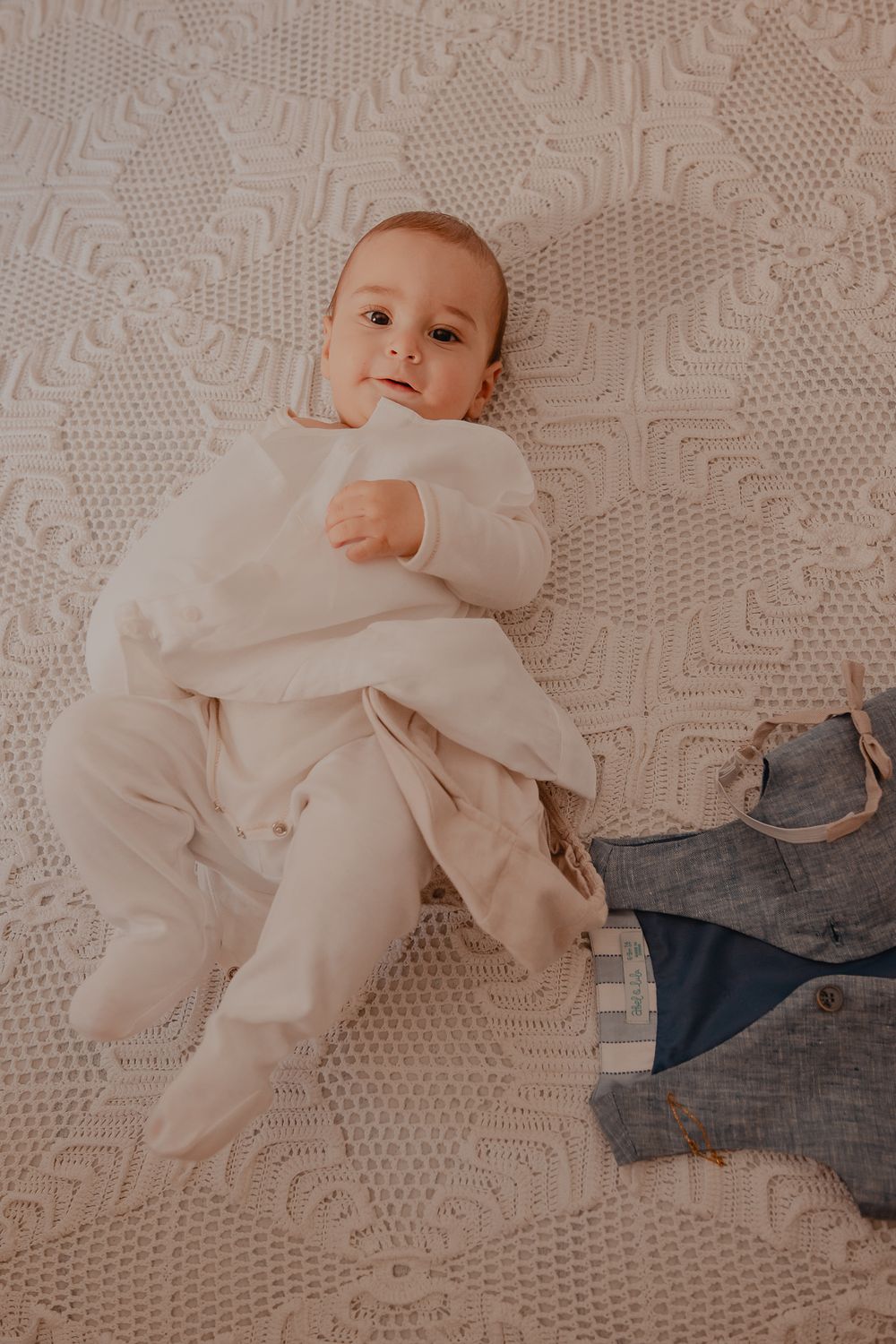 Baby in white outfit lying on textured white blanket with denim fabric visible.