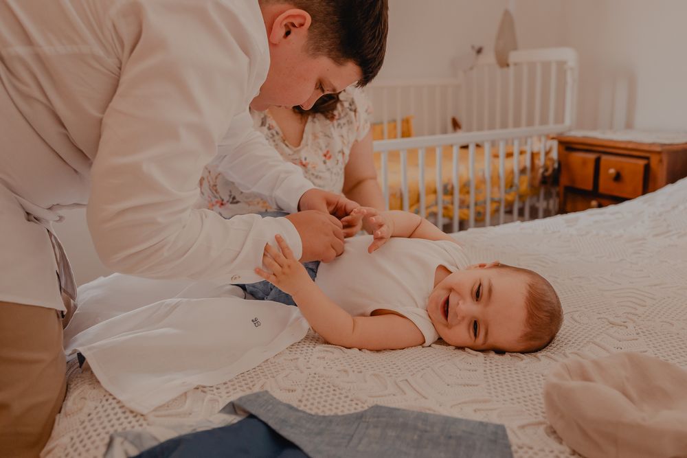 Baby in white clothing being dressed on textured bedding near wooden crib.