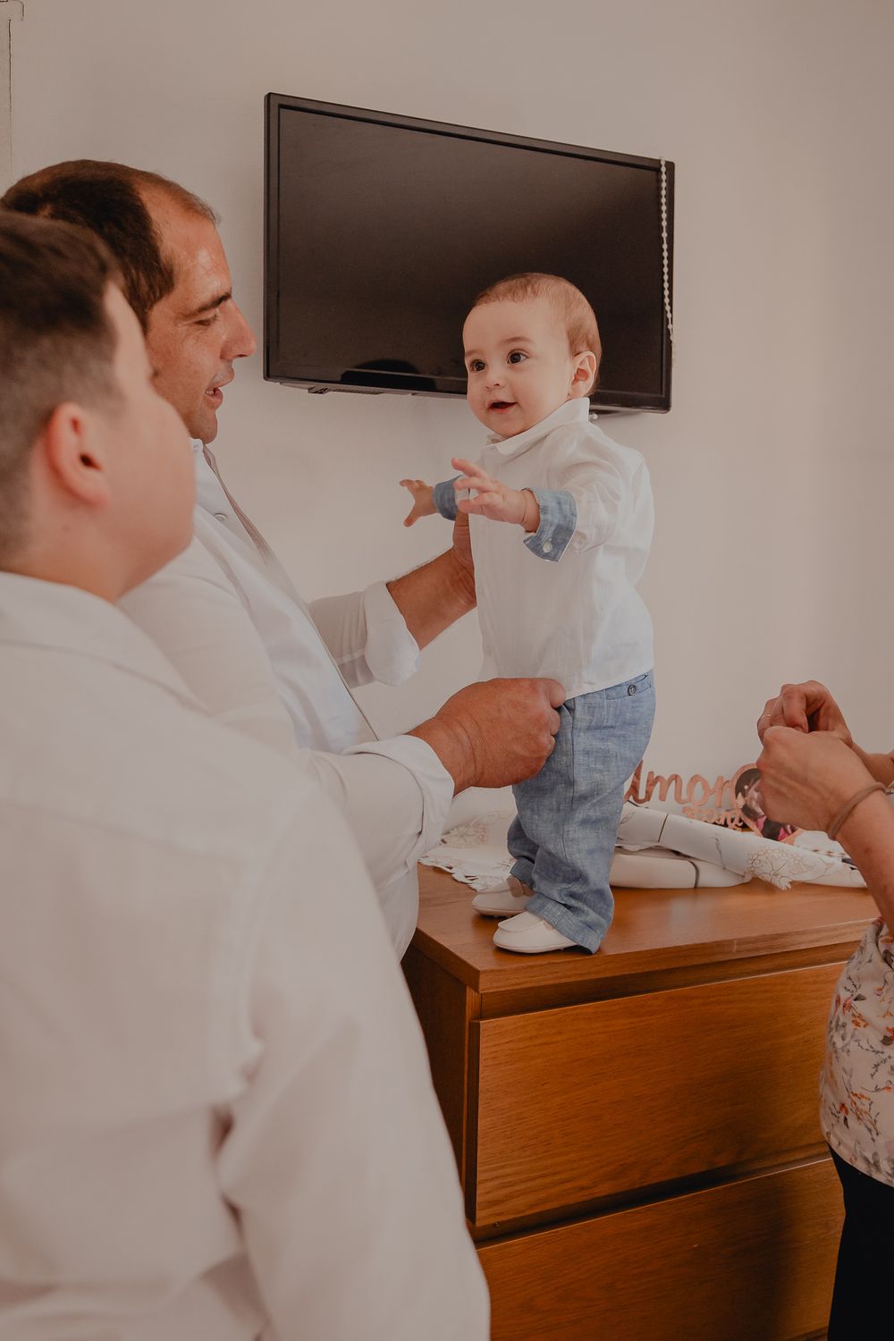 Baby standing on wooden dresser while interacting with adults.