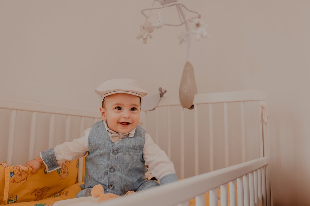 Baby in gray vest outfit sits in white crib with decorative wall art visible in background.