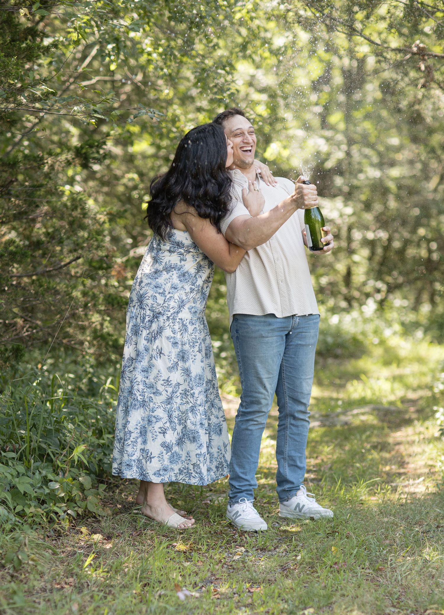 Two people celebrate outdoors with champagne in a sun-dappled wooded setting.