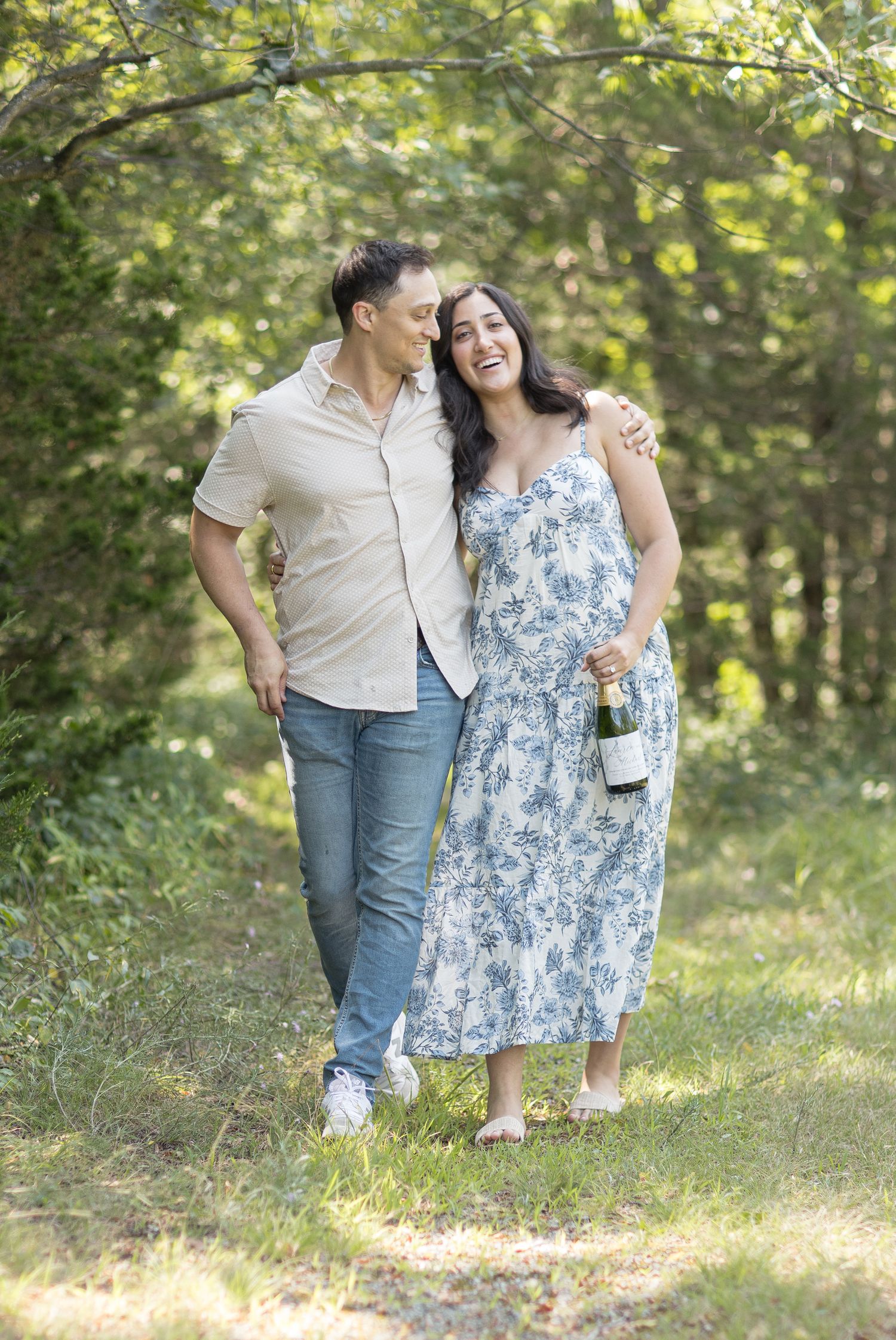 A couple enjoys a romantic outdoor photoshoot in casual attire with sunlit trees and greenery in the background.