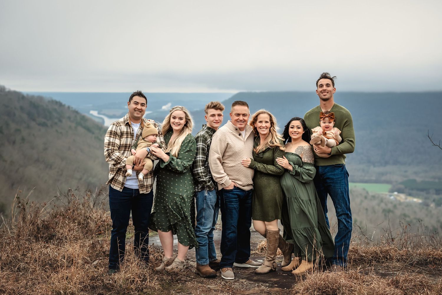 An extended family standing close together on a mountain overlook with a scenic valley and cloudy sky in the background.