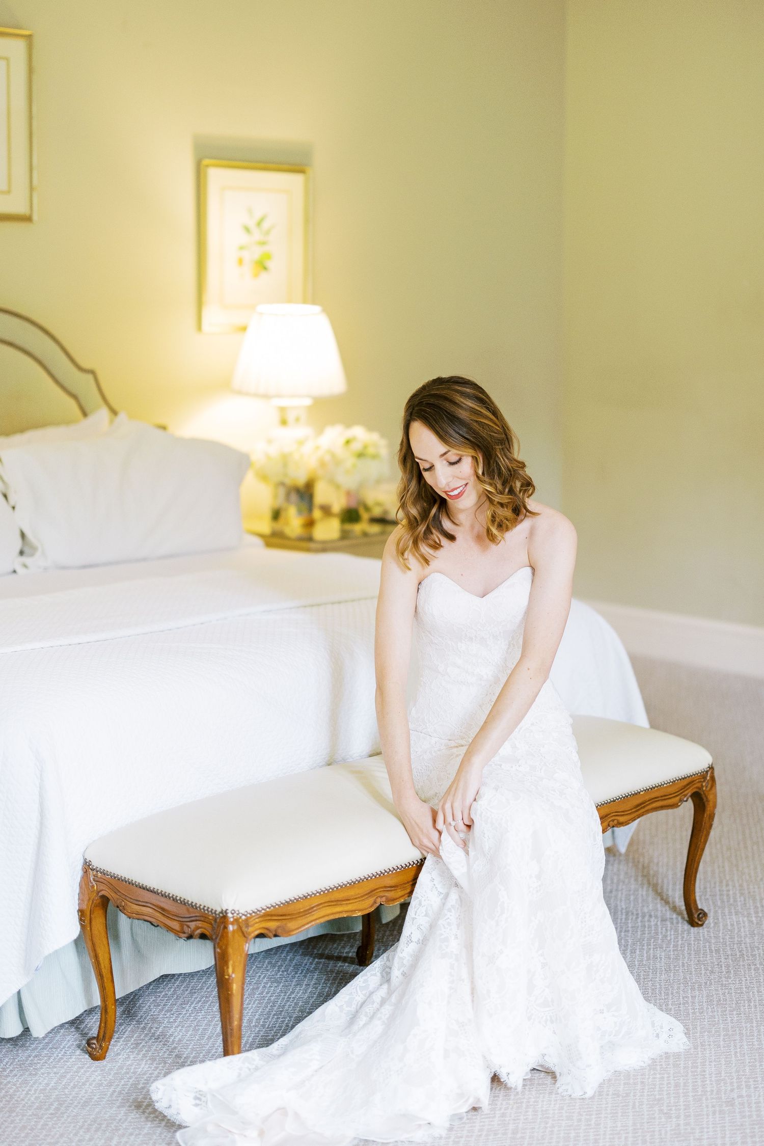 A bride in a flowing white gown sits elegantly on a wooden chair in a softly lit room with neutral walls.