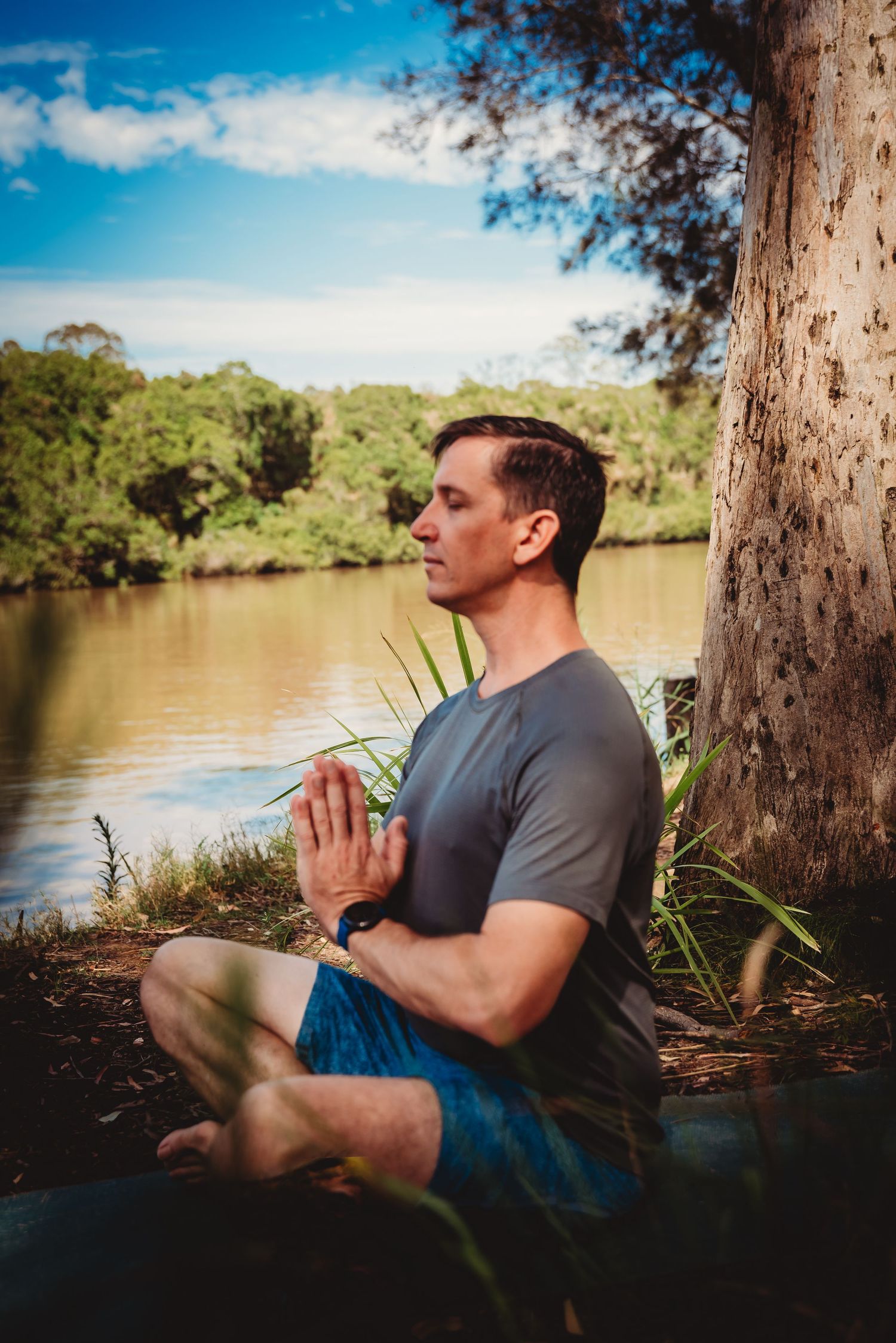 A person meditates in a cross-legged position beside a tranquil river on a sunny day in nature.