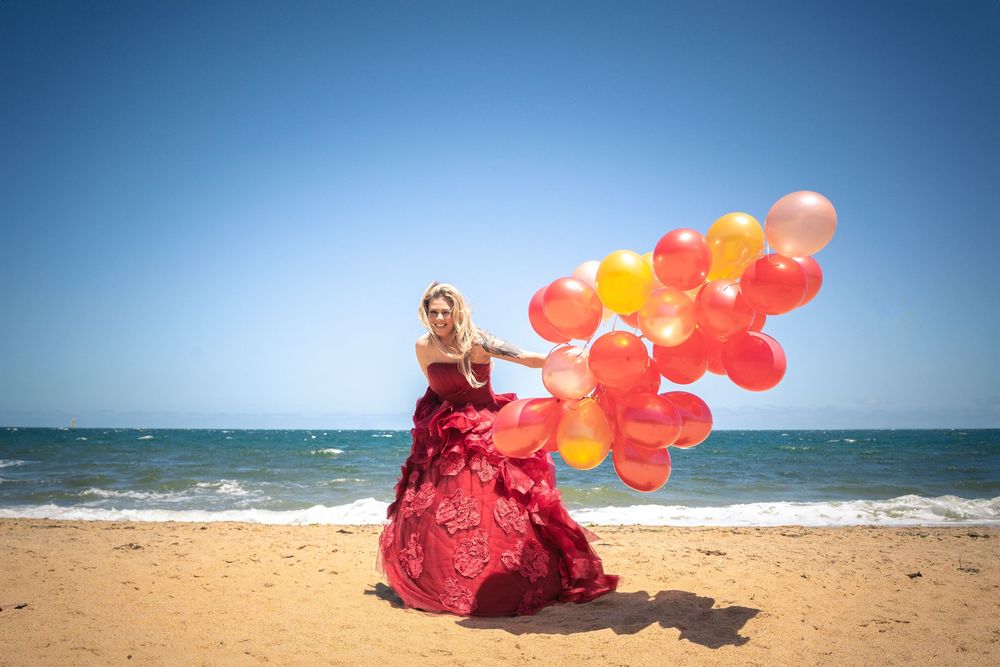 A person in a flowing red dress holds bright balloons on a sunny beach against a blue ocean backdrop.