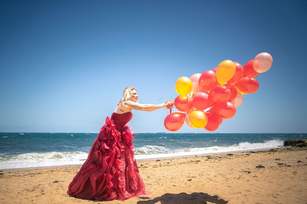 Figure in flowing red dress holds colorful balloons on sunny beach with ocean view in background.