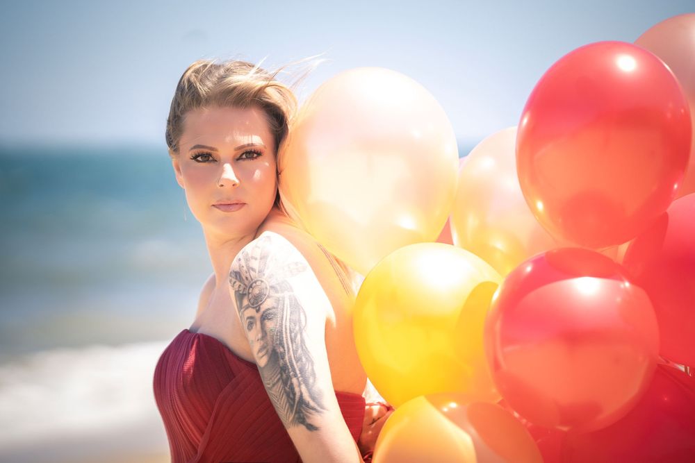 Person in red dress poses on beach with colorful balloons against blurred ocean backdrop.