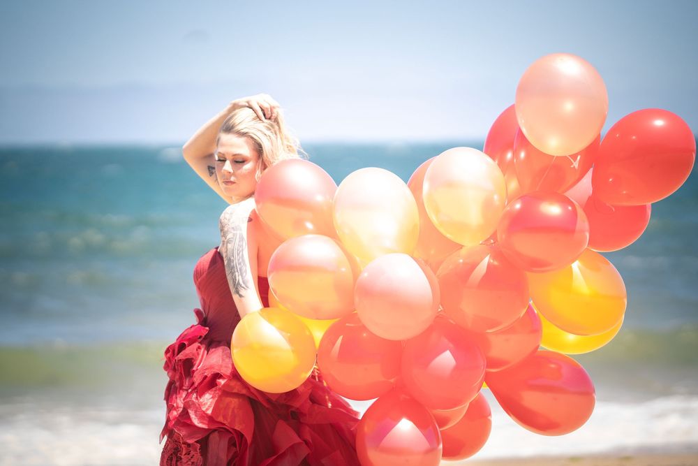 A figure in a red dress holds a bundle of orange and yellow balloons on a sunny beach with blue ocean backdrop.