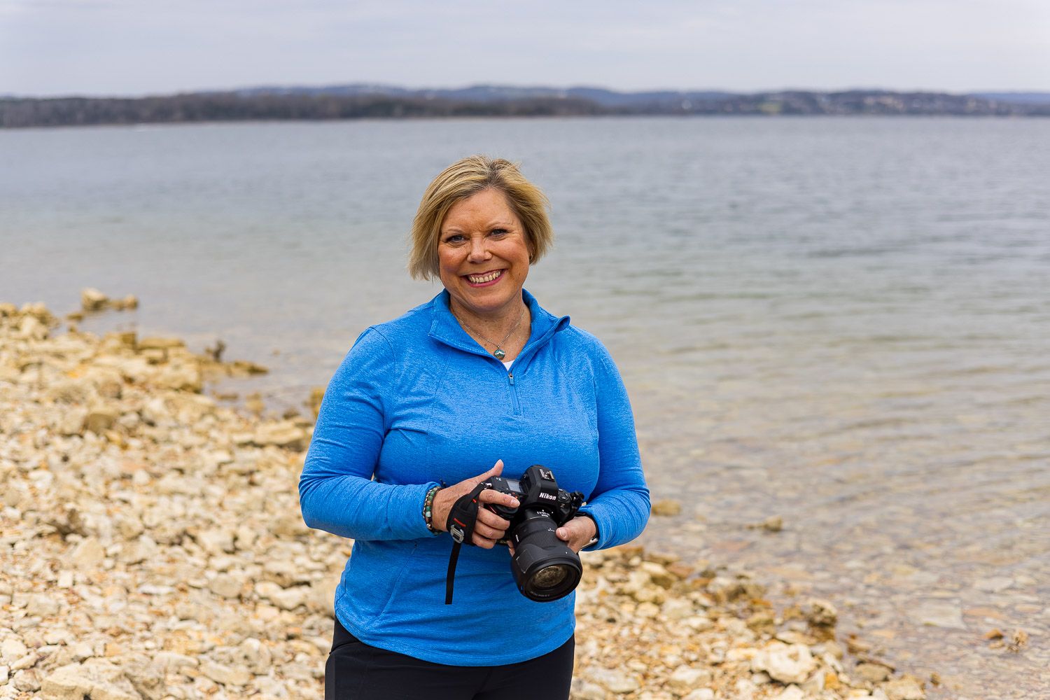 Smiling woman in blue jacket holds camera on rocky lakeside shore.