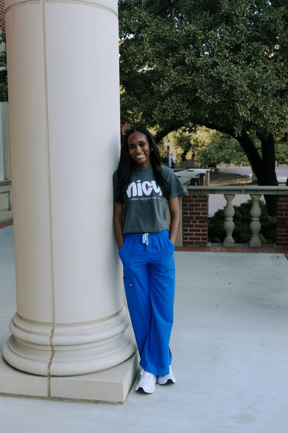 A person wearing a NICA t-shirt and blue sweatpants poses casually next to a white column on a brick building.