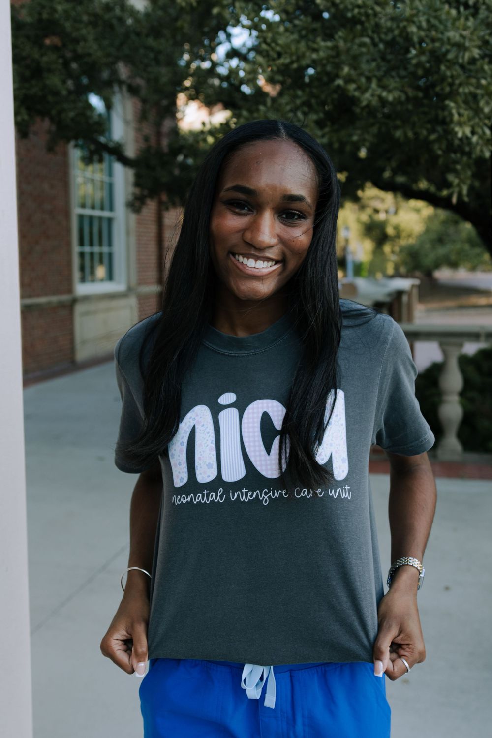 A person in a dark NICA t-shirt smiles while standing on a paved walkway with trees in the background.