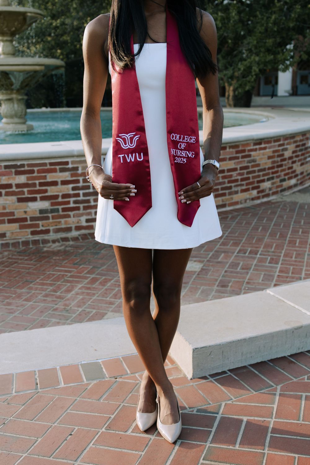 Close-up of white graduation dress with red TWU graduation stole and gold accessories on brick courtyard.