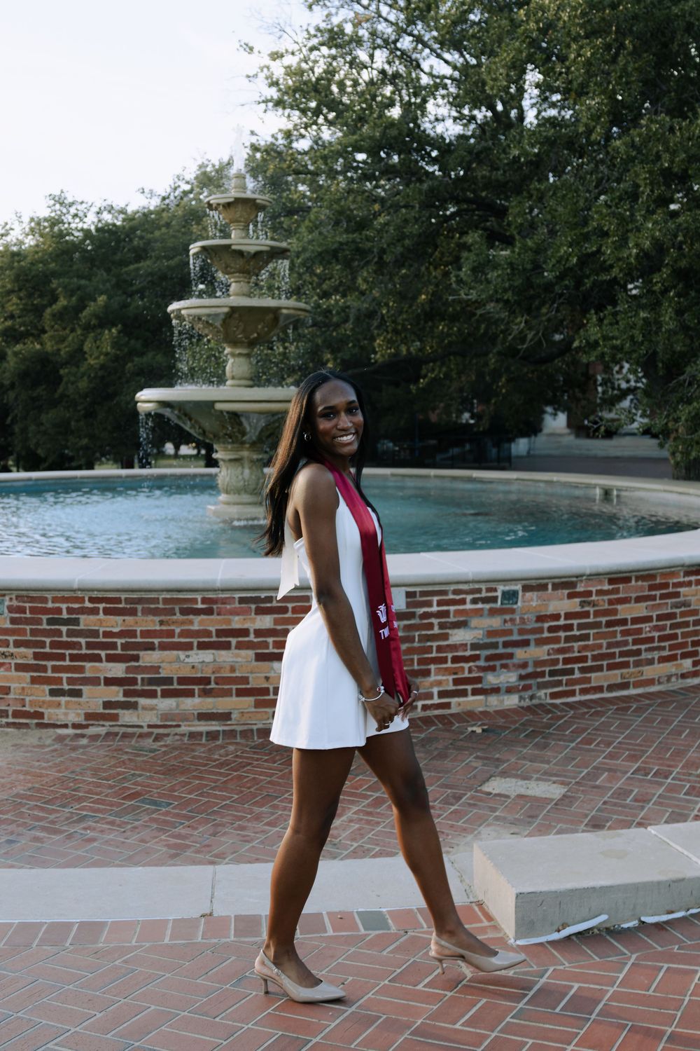 A graduate poses in a white dress and graduation gown on a brick patio near a fountain.