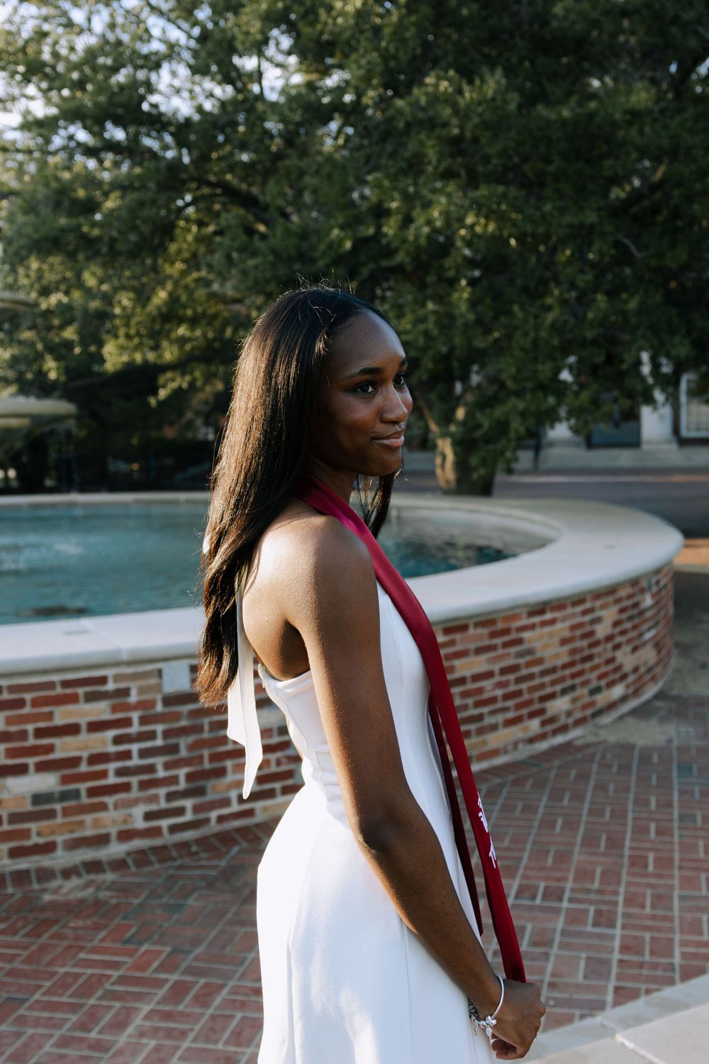 A person in a white dress and red stole poses by a curved brick fountain wall during graduation.