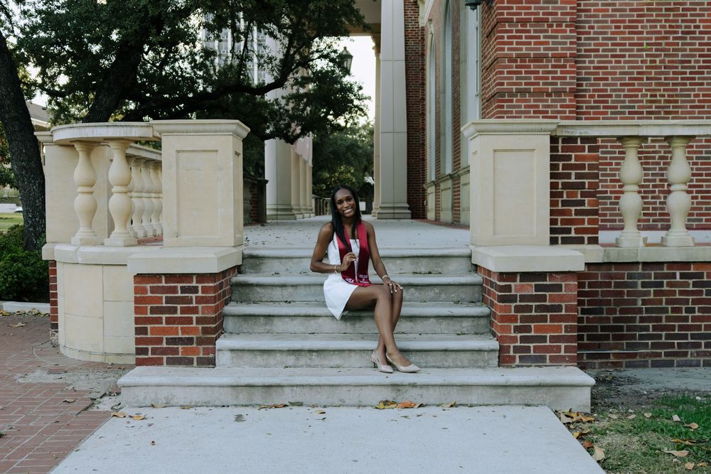 Person in a burgundy blazer and shorts sits on concrete steps of a brick building with white columns.