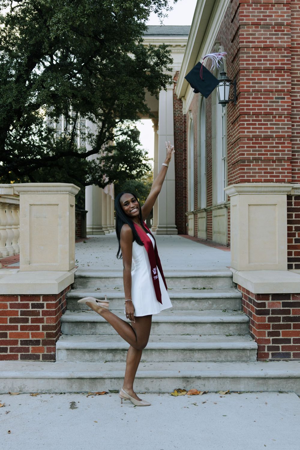 Graduate in white dress and red stole poses on brick steps of a campus building.