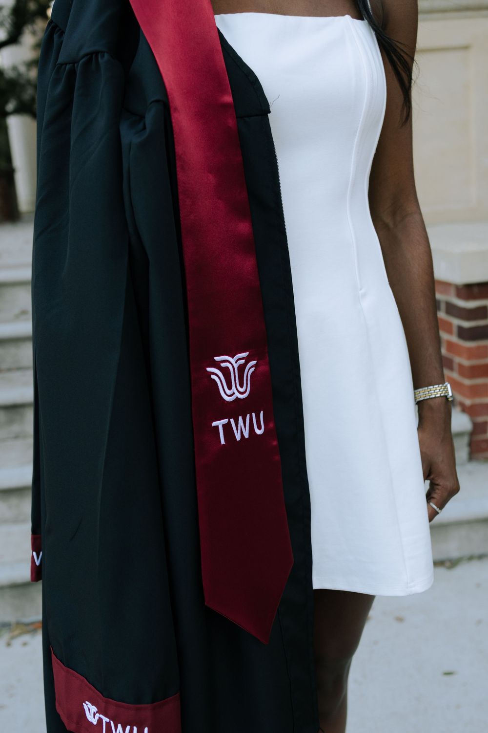 A graduate wearing a white dress and TWU graduation stole stands for photos outdoors.