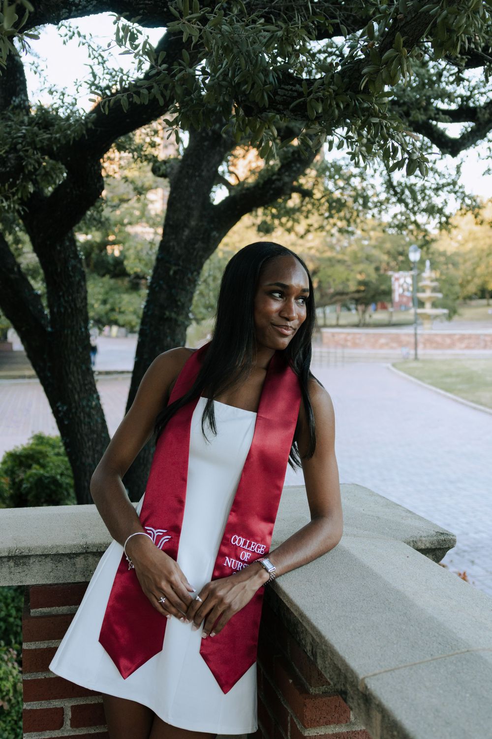 Graduate in white dress and red stole poses elegantly among campus trees.