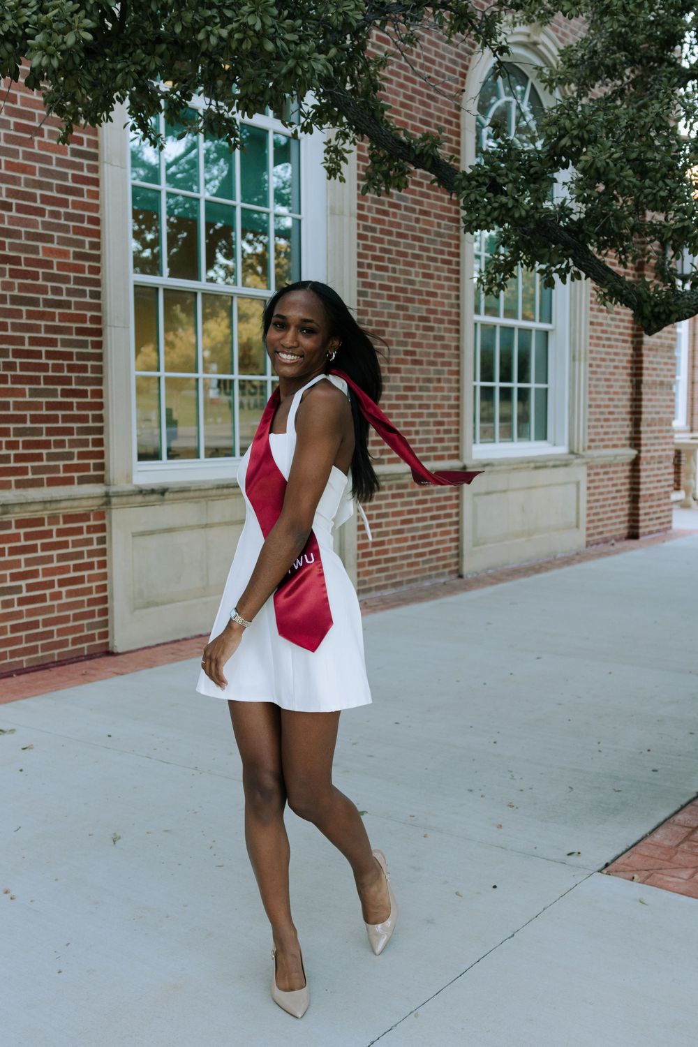 A person in a red and white sleeveless dress walks along a brick building sidewalk wearing nude heels.