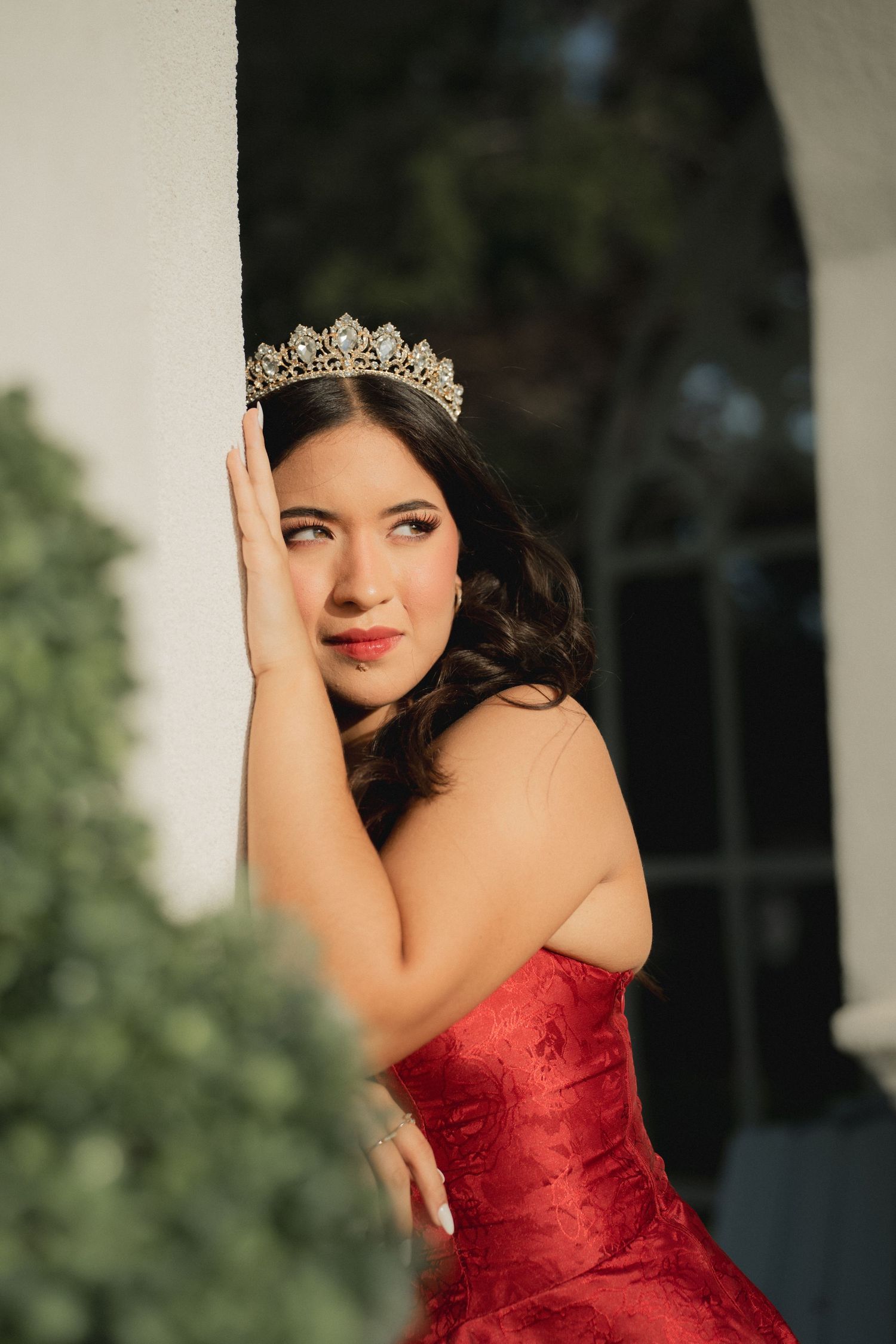 A glamorous photoshoot featuring a red formal gown and tiara against a white column backdrop.