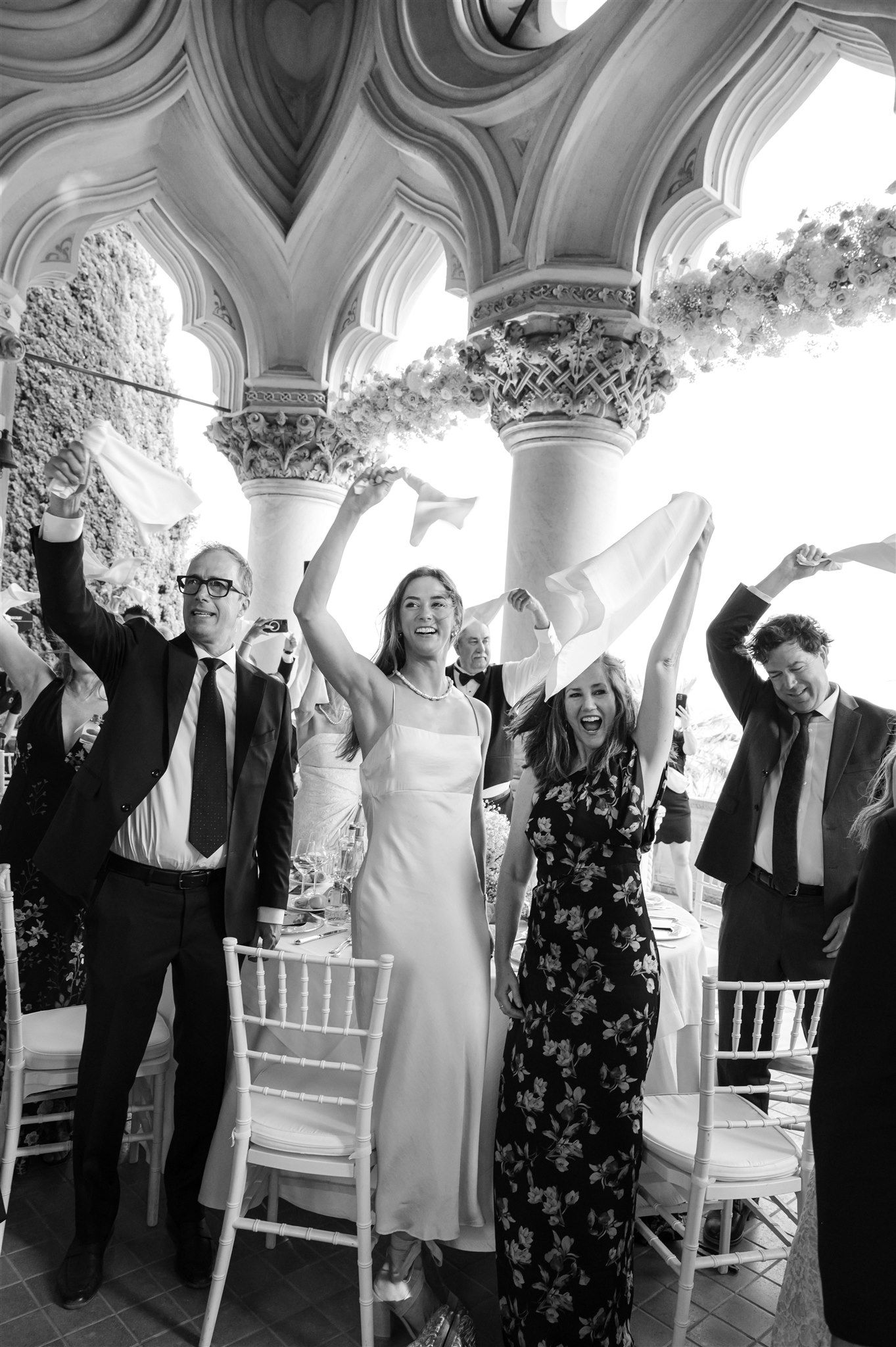 Wedding guests celebrate joyfully under ornate gothic arches in black and white photography.