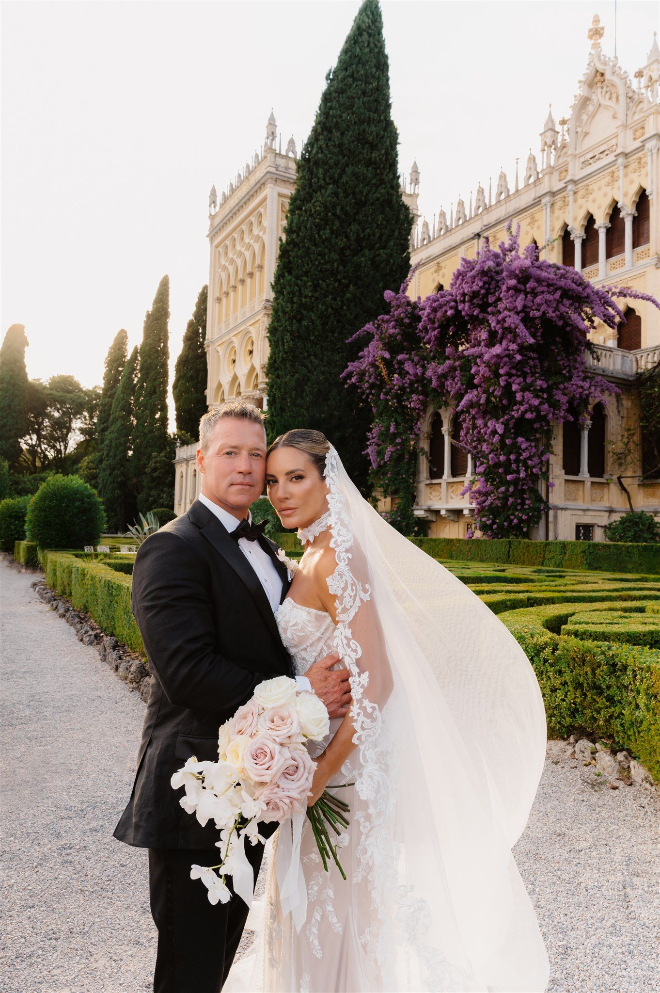 Wedding couple poses in front of ornate villa with blooming purple bougainvillea and manicured gardens.