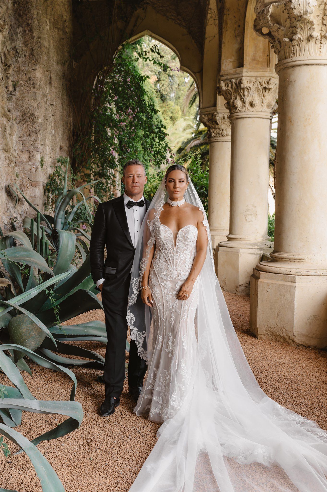 Wedding couple stands under arched colonnade with trailing plants and ornate stone columns.